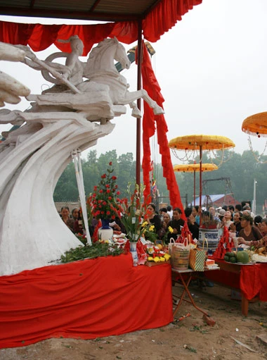 Từ sáng sớm, hàng nghìn người đã đổ về khu đúc tượng cạnh Học viện Phật giáo VN (Sóc Sơn, Hà Nội). Theo lịch âm, hôm nay là ngày "trùng cửu", ngày có ý nghĩa thiêng liêng đặc biệt, rất thích hợp để tiến hành đúc tượng Thánh Gióng - Đức Thánh Phù Đổng Thiên vương. Một mô hình thu nhỏ đặt cạnh khu đúc tượng để phật tử làm lễ, chiêm bái. Khi hoàn thành, tượng cao gần 11 m, độ vươn chéo lên trời khoảng 16 m. Rất nhiều người góp trang sức bằng kim loại quý để hòa cùng dòng đồng nóng chảy lúc đổ vào khuôn. Tại khu đất được cách ly cạnh đó, hai chiếc khuôn của phần đầu ngựa và thân trên Thánh Gióng đang gấp rút được hoàn tất những khâu cuối cùng. Đây là 2 thớt đầu tiên được đúc của tượng đài. Một số chi tiết khác của tượng đài cũng được làm trong mẻ đầu tiên. Hàng trăm thanh đồng đỏ nguyên chất được nung chảy trong lò ga hơn 1.000 độ C. Theo các nghệ nhân, thời gian từ lúc nấu đến lúc đổ đồng mất hơn 2 giờ. Người nghệ nhân cẩn thận trát kín mạch và tạo miệng khuôn để đổ đồng nóng chảy vào. Sau 2 giờ nung, mẻ đồng nóng chảy đổ ra... ...trong khi các nghệ nhân kính cẩn thắp hương mong mọi việc suôn sẻ. Bên ngoài hàng rào, hàng nghìn người chen lấn mong được vào chiêm ngưỡng cận cảnh. Tới dự buổi lễ sáng nay có nguyên Tổng bí thư Đỗ Mười, cùng nhiều vị lãnh đạo. Ngay trước thời khắc 9h9, hòa thượng Thích Thanh Tứ, Phó chủ tịch Trung ương hội Phật giáo VN(người đeo tràng hạt) cùng nhiều quan chức tiến vào khu đúc tượng. Hàng trăm món đồ kim loại quý được thả vào khuôn... ... trước khi dòng đồng đỏ nóng chảy được rót. Theo các nghệ nhân, công đoạn khó nhất của quá trình đúc tượng là khâu làm khuôn: mất 2 tháng. Khuôn phải làm sao để toát lên được thần thái của Thánh Gióng và toàn bộ hoa văn, họa tiết tinh xảo của tượng với đúng tỉ lệ 1:1. Sau khi đổ đầy đồng vào khuôn, cần tới 4 ngày để làm nguội tự nhiên rồi mới phá khuôn.